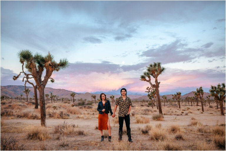Couple holding hands in Joshua Tree desert with sunrise and moon behind them