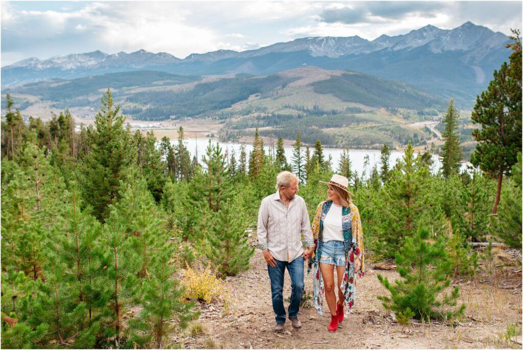 Couple walks holding hands through forest with lake and mountains in background