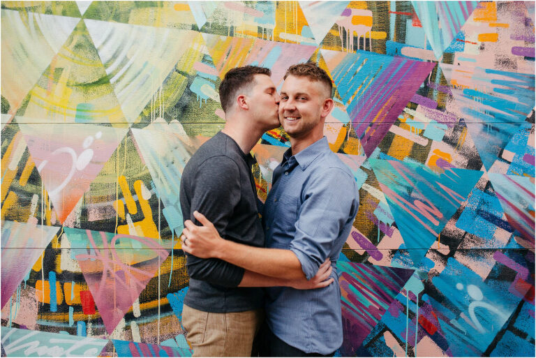 A couple embracing with one man giving the other a kiss on the cheek with bright street art behind them